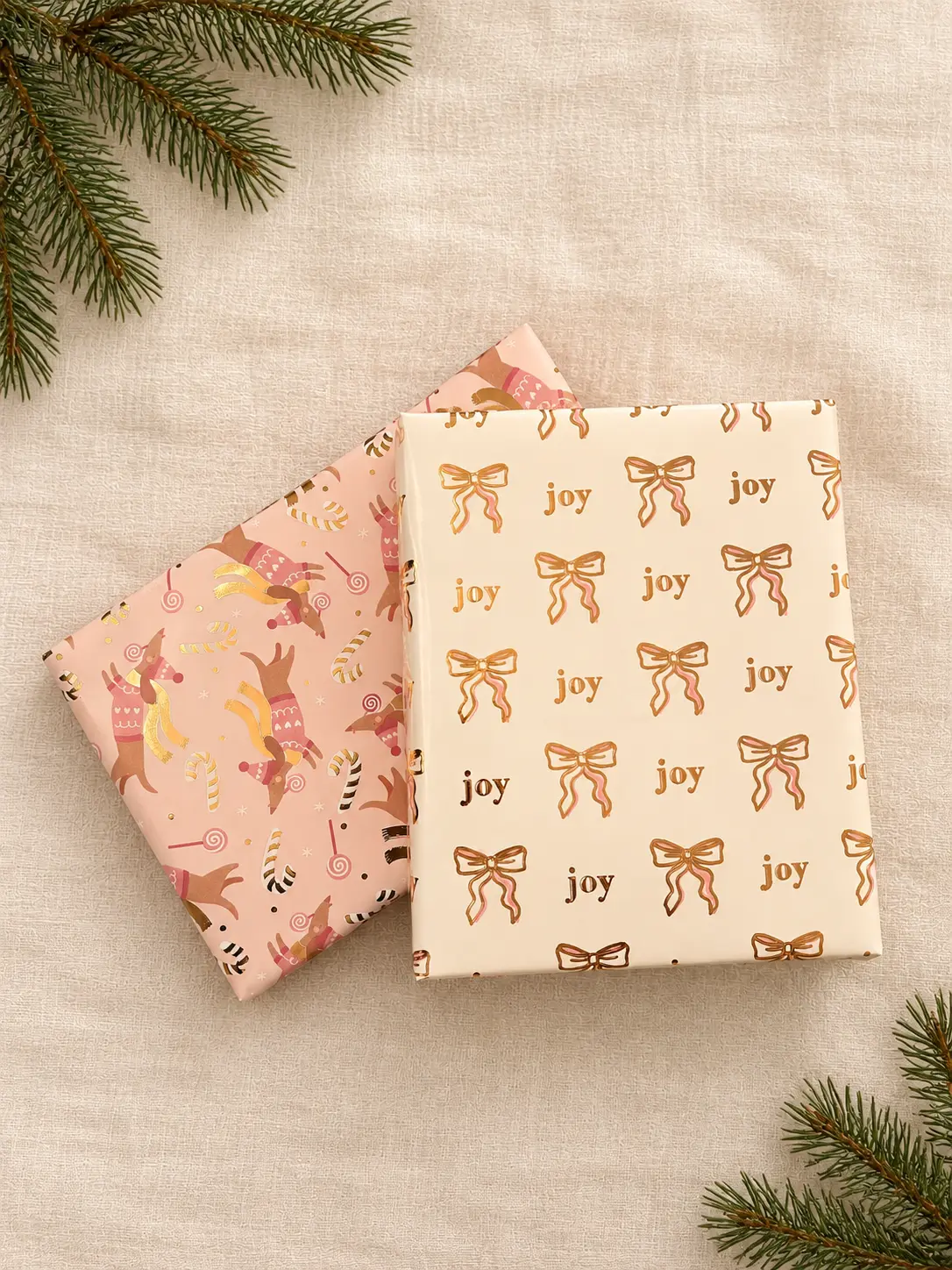 Two holiday gifts stacked on ivory linen, one wrapped in cream paper printed with gold ribbon bows and the word joy, the other in pink paper printed with dachshunds in winter sweaters